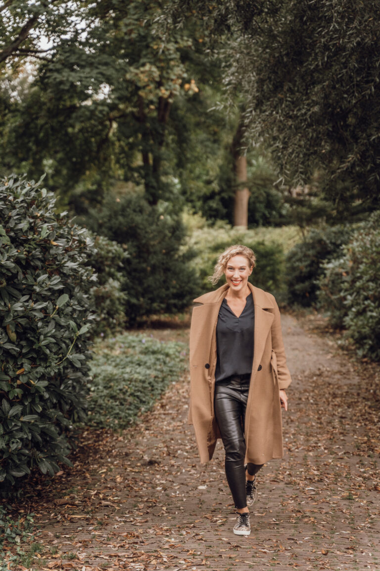Blonde vrouw met lange bruine jas wandelend met sneakers aan komt lachend in beeld.