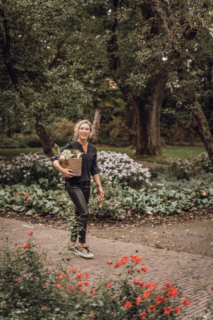 Blonde vrouw met krullen draagt een mand met groenten onder haar arm in pad met prachtige bloemen.