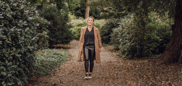Blonde vrouw met lange bruine jas wandelend met sneakers aan komt lachend in beeld. park met bomen en veel groen.