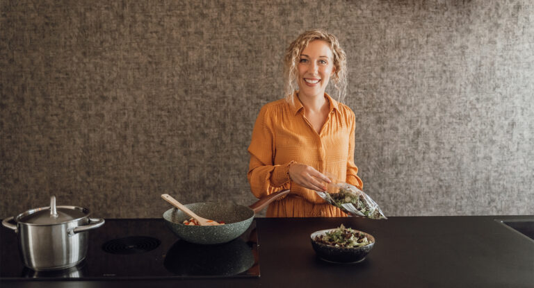 Blije vrouw maakt een gezonde salade in een moderne donker kleurige keuken.