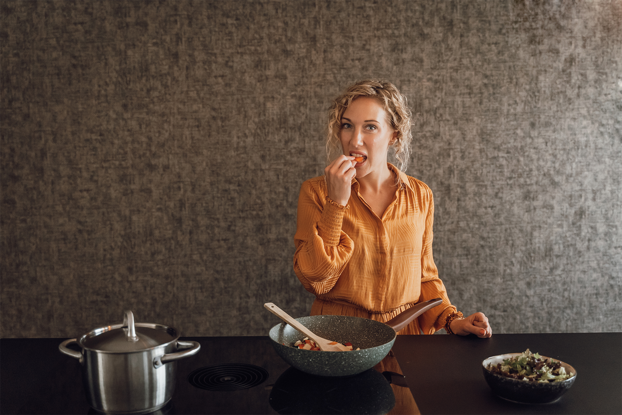 Vrouw met blonde krullen is een gezonde maaltijd aan het voorbereiden. Ze eet een groente uit de pan, voor haar links staat een salade met een mix sla.