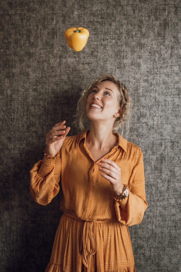 Vrouw met blonde krullen gooit speels een gele paprika in de lucht.