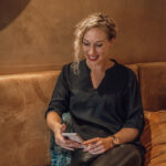 Blonde vrouw met krullen met glimlach en telefoon in de hand.