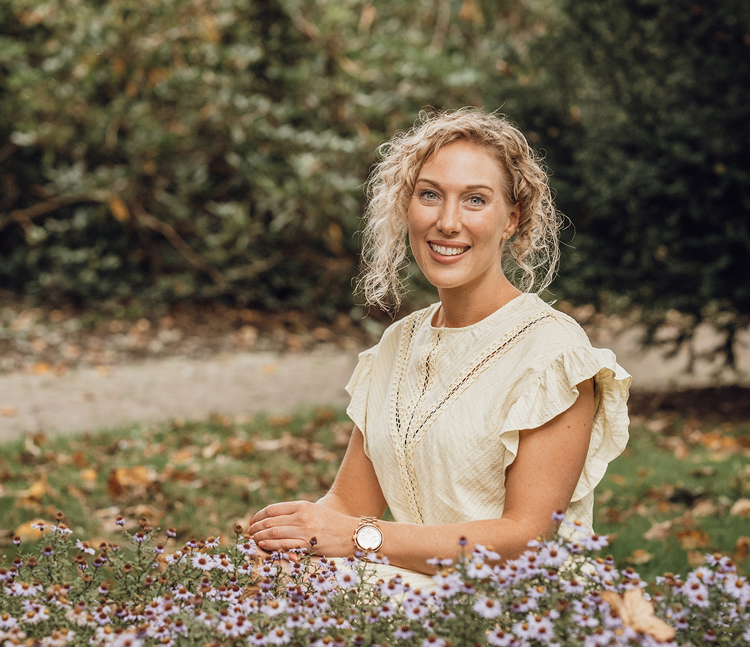 Blonde vrouw met krullen zit stralend voor een veld met bloemen.