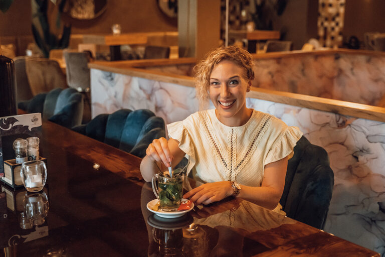 Vrouw glimlachend met blonde krullen drinkt een kruidenthee.