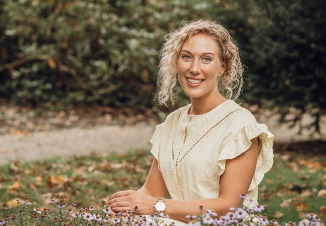 Blonde vrouw met krullen zit stralend voor een veld met bloemen.